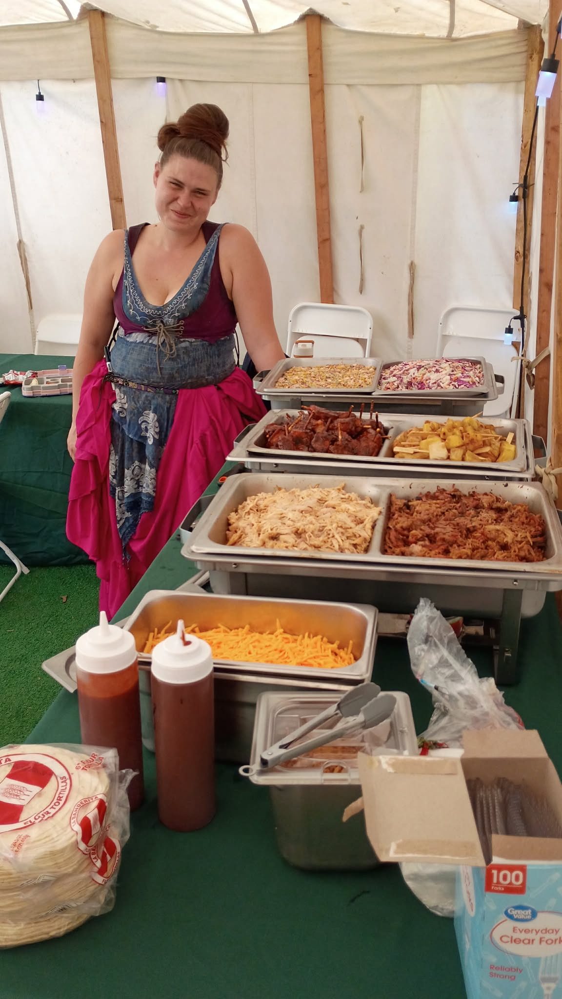 A woman in a renaissance faire dress smiling beside a buffet of chafing dishes holding pulled meats, glazed bites, grilled pineapple, coleslaw, and corn salad, with tortillas, cheese, and sauces in front
